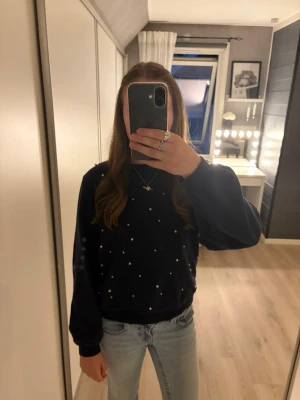 Mörkblå sweatshirt med pärldetaljer - Mysig mörkblå sweatshirt med pärldetaljer framtill. Tröjan har rund halsringning, långa ärmar och ribbade muddar vid ärmslut och nederkant. 
