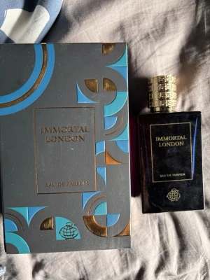 Immortal London Eau de Parfum - Parfymen Immortal London Eau de Parfum kommer i en elegant mörk flaska med gulddetaljer och matchande förpackning i svart, blått och guld. Flaskan har en fyrkantig form och ett dekorativt guldlock som ger en lyxig känsla. Perfekt för dig som gillar exklusiva dofter.