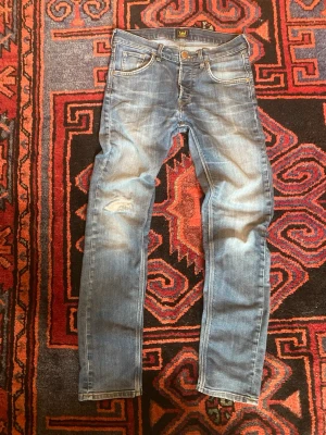 Blå slitna jeans från Lee - Säljer ett par ljusblå jeans från Lee med slitningar och tvättade detaljer. Modellen har smal passform, klassiska fem fickor och knappgylf. Perfekta för en avslappnad och trendig look.