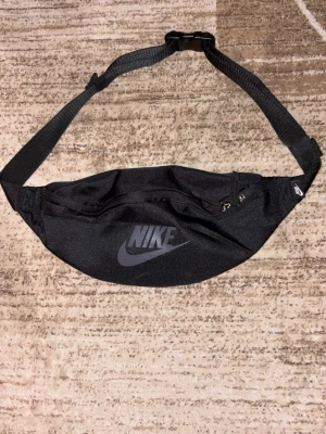 Svart magväska från Nike - Snygg svart magväska från Nike med justerbart band och spänne. Väskan har två dragkedjefack, ett fram och ett bak, samt stor Nike-logga på framsidan. Perfekt för att bära det viktigaste när du är på språng.