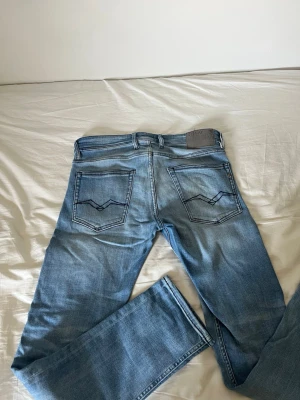 Ljusblå jeans från Replay Anbas  - Säljer ett par ljusblå jeans från Replay med raka ben och klassiska femficksdetaljer. Jeansen har tydliga kontrastsömmar på bakfickorna och en Replay-lapp i läder bak i midjan. Perfekta för en avslappnad och trendig stil.