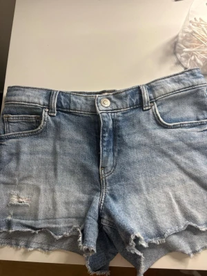 Blå jeansshorts från ONLY - Säljer ett par ljusblå jeansshorts från ONLY med råa kanter och slitningar. Klassisk femficksmodell med knapp och dragkedja framtill. Perfekta för sommaren och ger en avslappnad vibe till din outfit. passar även folk som bär storlek XS/S