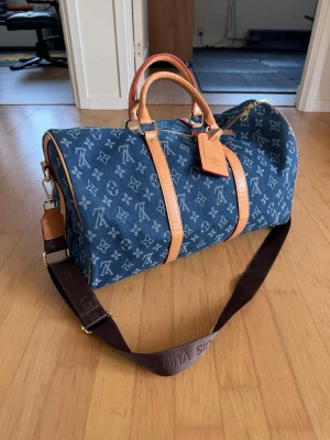 Louis Vuitton dufflebag - Snygg blå dufflebag i denim-look med klassiskt monogrammönster och detaljer i ljusbrunt läder. Väskan har både handtag och en bred, justerbar axelrem i mörkbrunt tyg. Perfekt för dig som vill ha en trendig och rymlig väska med exklusiv känsla.