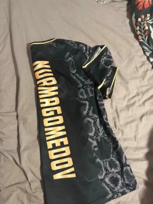 UFC t-shirt med Nurmagomedov-tryck - Säljer en svart UFC t-shirt med Kurmagomedov-tryck i gult på ryggen och coolt mönster framtill. T-shirten har gula detaljer vid ärmar och hals, samt tryck från crypto.com och UFC-logga. Perfekt för dig som gillar MMA och sportiga plagg. Storlek S (LITEN I STORLEK)