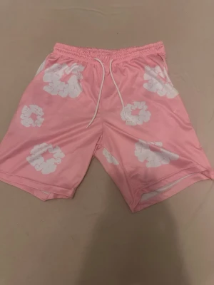 Rosa shorts med vita moln - Säljer ett par rosa shorts med vita molnliknande mönster. Shortsen har resår i midjan och vita dragsnören framtill. Tillverkade i ett mjukt och glansigt material som känns skönt mot huden. Perfekta för en avslappnad och trendig look.