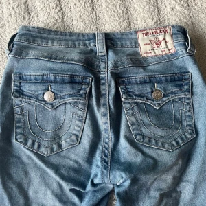 True Religion ljusblå bootcut jeans - Säljer ett par ljusblå jeans från True Religion, modell BECCA Mid Rise Bootcut. Jeansen har snygga kontrastsömmar, dekorativa bakfickor med lock och silverknappar. Klassisk bootcut passform och tillverkade i mjuk denim. Perfekta för en avslappnad men trendig look.