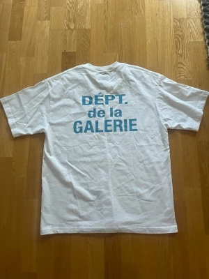 Vit t-shirt med blå texttryck - Vit t-shirt med blått tryck 'DÉPT. de la GALERIE' på både bröstet och ryggen. Klassisk passform med korta ärmar och rund hals. Perfekt för en avslappnad och trendig stil.