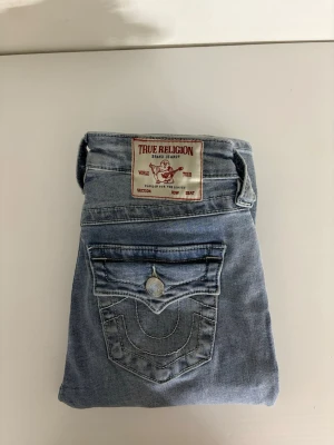 Ljusblå True Religion jeans - Säljer ett par snygga ljusblå jeans från True Religion med klassisk fickdesign och tydliga sömmar. Jeansen har en knappgylf och fem fickor, samt en patch med logga bak. Perfekta för en avslappnad och trendig stil.