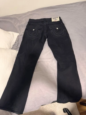 Svarta True Religion jeans - Säljer ett par svarta jeans från True Religion med raka ben och ikoniska kontrastsömmar på bakfickorna. Jeansen har två stora fickor bak med lock och silverfärgade knappar samt en patch med logga i midjan. Perfekta för en cool och avslappnad stil.