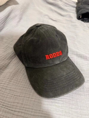 Bolaget keps - Snygg svart keps med böjd skärm och röd brodyr med texten 'RODEO' framtill. På sidan finns en liten vit brodyr med bokstäverna 'BR'. Kepsen är i klassisk baseballmodell och tillverkad i bomullstyg.