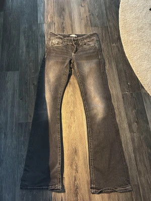 Grå bootcut jeans med kontrastsömmar - Säljer ett par grå bootcut jeans med tydliga vita kontrastsömmar och snygga fickdetaljer med knappar fram. Jeansen har en klassisk femficksmodell och är tillverkade i ett stretchigt denimtyg som sitter skönt. Perfekta för dig som gillar en avslappnad men trendig look. Aldrig använda 
