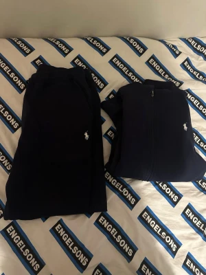 Mörkblå Polo Dress - Mysigt mörkblått loungewear-set från Ralph Lauren med matchande byxor och hoodie. Hoodien har dragkedja, fickor och dragsko i huvan. Både byxorna och tröjan har den klassiska Polo-loggan broderad på bröstet och benet. Perfekt för chill dagar hemma.