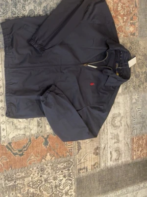 Mörkblå vindjacka från Polo Ralph Lauren - Snygg mörkblå vindjacka från Polo Ralph Lauren med dragkedja framtill och klassisk krage. Jackan har elastiska muddar vid ärmsluten och en liten röd logga broderad på bröstet. Perfekt för blåsiga dagar och enkel att matcha med olika outfits.