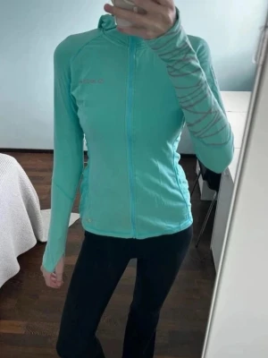 Define jacket - Snygg turkos träningsjacka från SOC med hel dragkedja framtill och hög krage. Jackan har långa ärmar med diskreta gråa detaljer på ena ärmen och är tillverkad i ett stretchigt funktionsmaterial som passar perfekt till träning eller löpning.