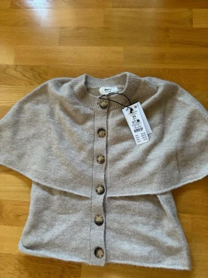 Beige kofta från Gina tricot med knappar - Snygg beige kofta från Gina tricot med rund hals och stora träknappar framtill. Koftan är stickad och har ribbade detaljer vid halsen. Perfekt att slänga över en t-shirt för en chill look.