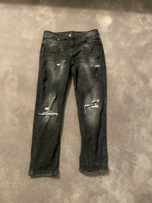 Svarta distressed jeans med slitningar - Coola svarta jeans med slitningar och paint splatter-effekt. De har klassisk femficksdesign, dragkedja och knapp framtill samt en extra zip-detalj på fickan. Jeansen är raka i modellen och gjorda i denim.