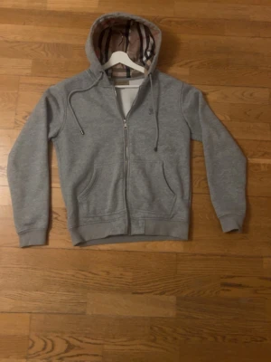 Grå Burberry Hoodie Small - Säljer en grå Burberry zip hoodie med klassiska rutmönstret på insidan. Sjukt skön i materialet och sitter riktigt bra. Perfekt nu till sommarkvällar när det blir lite kallare. Bra skick.