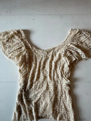 Beige spetsblus från Divided H&M - Söt beige blus från Divided by H&M med volangdetaljer och breda, korta ärmar. Tillverkad i spetsmaterial som ger en romantisk känsla. Perfekt för dig som gillar en feminin och trendig stil.