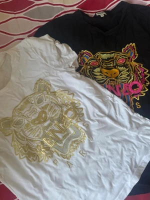 Kenzo t-shirts med tigermotiv - Två snygga Kenzo t-shirts med ikoniskt tigermotiv på bröstet. En är vit med guldfärgat broderi och den andra är svart med färgglatt broderi i gult, rosa och blått. Båda har rund hals och normal passform. Perfekta för att lyfta din outfit. 300 för båda 150/styck kan mötas upp Helsingborg o Malmö annars tillkommer frakt kostnad pris .