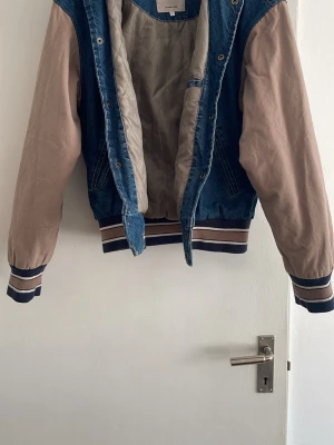 Jack & Jones jeansjacka med tryck - Snygg jeansjacka från Jack & Jones med blå denim på ryggen och beige ärmar. Stor vit text 'SOCIETY' på ryggen och randiga muddar i blått och beige. Jackan har quiltat foder och en avslappnad college-inspirerad look. Inget kvitto tyvärr.