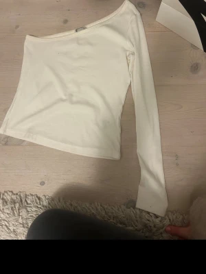 Vit oneshoulder topp i bomull - Trendig vit oneshoulder topp i mjukt bomullsmaterial. Toppen har lång ärm på ena sidan och är axelbandslös på andra, vilket ger en cool asymmetrisk look. Perfekt att styla med jeans eller kjol för en modern vibe. Tror i storlek S💓 Skriv för frågor eller fler bilder!