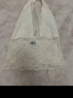 Vit spetsbralette från Gina Tricot - Säljer en vit bralette från Gina Tricot i storlek M. Den är gjord i mjuk spets med blommönster och har halterneck-modell. Braletten har en liten rosett framtill och fina vågiga spetskanter. Perfekt för dig som gillar romantisk och feminin stil.