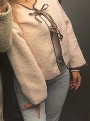 Ljusrosa cardiganjacka med rosett - Söt ljusrosa cardiganjacka med bruna kanter och dekorativa rosetter framtill. Jackan har en boxy passform och är gjord i ett mjukt, fluffigt material. Perfekt för dig som gillar en gullig och trendig stil.