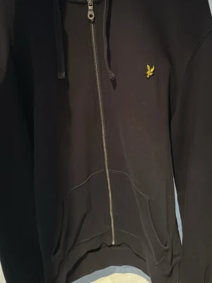 Svart zip-hoodie från Lyle & Scott - Svart hoodie med dragkedja från Lyle & Scott, prydd med den klassiska gula örnloggan på bröstet. Modellen har huva med snören och stora fickor framtill. Materialet är mjukt och bekvämt, perfekt för en chill look.