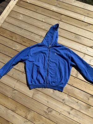 Blå zip-hoodie från Karl Kani - Blå hoodie med dragkedja från Karl Kani. Klassisk design med huva, ribbade muddar och två stora fickor framtill. Materialet är mjukt och bekvämt, perfekt för en avslappnad look. Diskret Karl Kani-logga broderad på bröstet.