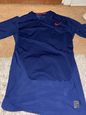 Blå Nike Pro långärmad träningströja - Nike Pro långärmad träningströja i blått med röd Nike-logga på bröstet. Tillverkad i Dri-FIT-material som andas och håller dig torr under intensiva pass. Tröjan har en sportig passform och rund hals, perfekt för gym eller löpning.