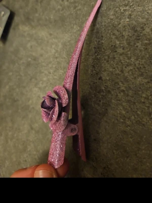 Glittrig lila hårklämma med ros - En snygg hårklämma i lila plast med glitter och en dekorativ ros på sidan. Perfekt för att ge frisyren en extra touch och hålla håret på plats. Klämman har en smal och avlång form med blommigt motiv som sticker ut.