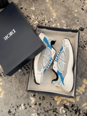 D***r sneakers i blått och vitt - Snygga sneakers från D***r  med coola-detaljer på sidan och hälen. Skorna har en mix av vitt, blått, grått och silver med svarta inslag. Låg modell med mesh och läderpaneler, perfekt för dig som vill sticka ut med exklusiva skor.
