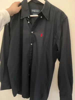 Mörkblå skjorta från Ralph Lauren - Säljer en mörkblå skjorta från Ralph Lauren med klassisk passform och knappar framtill. Skjortan är i 100% bomull och har det ikoniska röda Polo-logotypen broderad på bröstet. Perfekt för dig som gillar stilrena plagg med premiumkänsla.