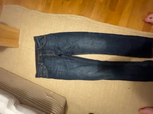 Mörkblå nudie jeans - Säljer ett par mörkblå slim fit jeans med klassisk femficksdesign och snygga slitningar på framsidan och baksidan. Jeansen har raka ben och är tillverkade i denim med en skön stretch. Perfekta för dig som gillar en modern och stilren look. Modellen är grim tim strl 32/32
