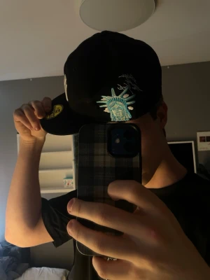 Svart New Era keps med NY-logga - Säljer en svart New Era keps med klassisk New York Yankees-logga i vitt framtill och broderad Frihetsgudinnan på sidan. Kepsen har platt skärm och sticker ut med coola detaljer. Perfekt för dig som vill ha en streetstyle-look. Storlek är 57.7 cm  eller 7 1/4