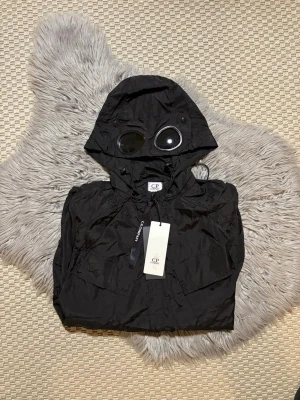 Svart vindjacka från C.P. Company - Snygg svart vindjacka från C.P. Company med ikonisk huva utrustad med inbyggda goggles. Jackan har två stora bröstfickor och justerbar dragsko i huvan. Tillverkad i lätt, glansigt material som ger en modern look. Perfekt för dig som vill sticka ut.