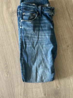 Blåa jeans med klassisk passform - Säljer ett par blåa jeans med klassisk femficksdesign och raka ben. Jeansen har en snygg tvätt med lätt slitning och är tillverkade i denim. Perfekt för dig som gillar en tidlös och enkel stil.