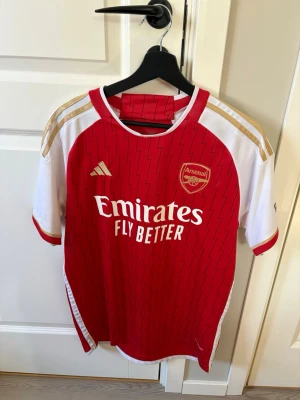 Arsenal matchtröja Adidas röd/vit - Säljer en officiell Arsenal matchtröja från Adidas i rött och vitt med gulddetaljer. Tröjan har klubbmärke och Adidas-logga på bröstet, samt Emirates Fly Better-tryck. Tillverkad i lätt och ventilerande material, perfekt för fotboll eller träning.