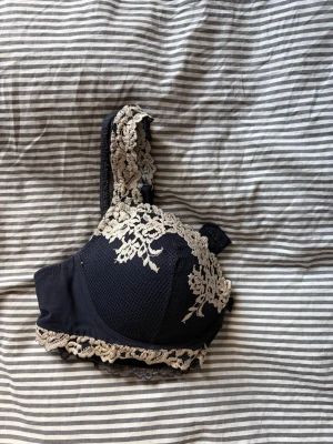 Svart intimissimi spets-bh med beige detaljer 75- C - En marinblå bh med vadderade kupor och dekorativa vita spetsdetaljer längs kanterna och axelbanden. Bh:n har justerbara axelband och hakknäppning bak. Materialet är en kombination av mjuk spets och slät textil för en elegant look.