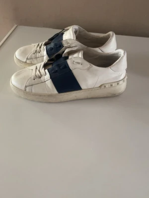 Valentino vita sneakers med blå detalj - Säljer ett par vita sneakers från Valentino med bred blå remsa över mitten och coola nitar på hälen. Skorna har klassisk låg profil, vita snören och är tillverkade i läder. Perfekta för dig som gillar stilrena men ändå unika sneakers, behöver en tvätt med händer och du har helt nya skor inga skador in eller ut ifrån