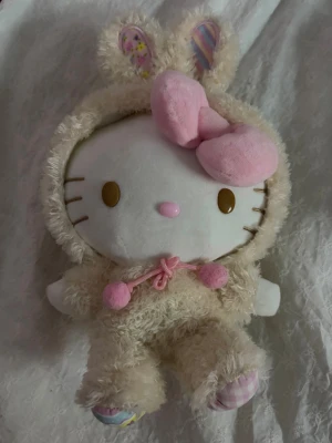 Hello Kitty gosedjur - Söt Hello Kitty plushie, klädd i fluffig beige kanindräkt med rosa detaljer och öron med mönster. Ansiktet är vitt med bruna ögon och rosa rosett.