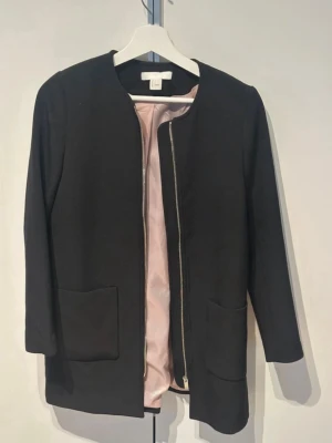 Svart tunn  kappa från H&M - Stilren svart kappa från H&M med rak passform och rund halsringning. Kappan har två stora fickor framtill och en silverfärgad dragkedja. Insidan är fodrad med ljusrosa tyg. Perfekt för dig som gillar enkel och modern design.