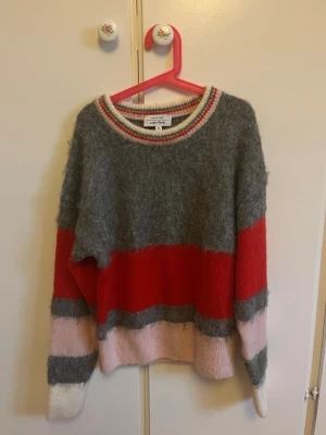Färgglad stickad tröja från & Other Stories - Säljer en stickad tröja från & Other Stories Paris Atelier med breda färgblock i grått, rött, rosa och vitt. Tröjan har rund hals med randig kant och långa ärmar. Materialet är mjukt och fluffigt, perfekt för att hålla dig varm under kyliga dagar.