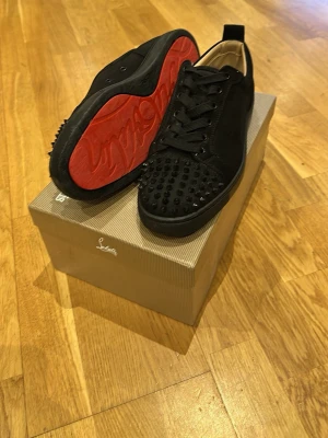 Svarta Louboutin sneakers med nitar - Säljer ett par svarta sneakers från Christian Louboutin med svarta nitar på tån och den klassiska röda sulan. Skorna har snörning, låg profil och är tillverkade i mocka och läder. Perfekta för dig som vill sticka ut med en lyxig och edgy stil.