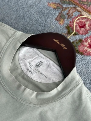 Brunello Cucinelli T-shirt  - Stilren ljusgrå t-shirt från Brunello Cucinelli med texten 'Simplicity in Elegance' tryckt framtill. Klassisk rund halsringning och korta ärmar. Slim fit-modell i mjuk tjock bomull som känns riktigt lyxig och skön. Nypris 4500kr, perfekt skick utom att tvättlappen är bortsprättad.
