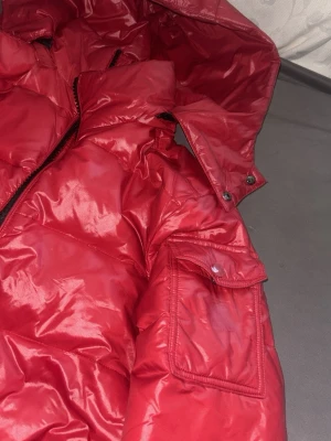 Röd pufferjacka med huva - Snygg röd pufferjacka med glansig finish och stor huva. Jackan har flera fickor med dragkedja och en extra ficka med lock på ärmen. Perfekt för kalla vinterdagar och riktigt varm. Stängs med dragkedja framtill.