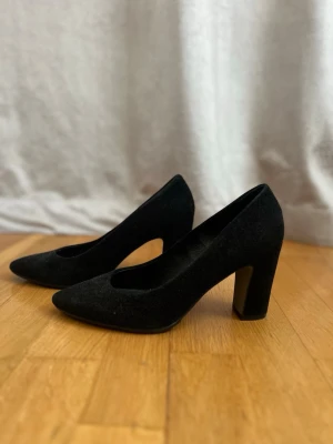 Svarta pumps med klack  - Stilrena svarta pumps från Clou med spetsig tå och bred klack. Skorna är tillverkade i mocka som ger en lyxig känsla. Perfekta för dig som vill ha en klassisk och elegant look.