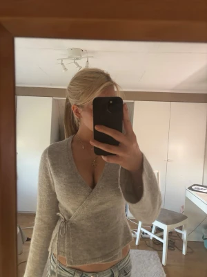 Grå omlott tröja från gina - Mysig grå omlottkofta med knytband i midjan och v-ringad hals. Koftan har långa ärmar och är tillverkad i ett mjukt, fluffigt material som ger en avslappnad och trendig look. 