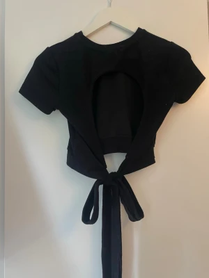Svart croppad topp med knyt från Gina - Snygg svart croppad topp med korta ärmar och öppen rygg som knyts baktill. Perfekt för dig som vill ha en trendig och edgy look. Toppen har en tight passform och är tillverkad i mjukt material som känns skönt mot huden. Strl M men sitter som en S, den e ganska kort. 