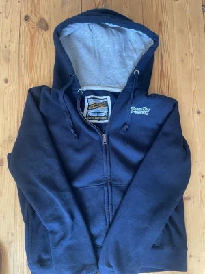 Mörkblå hoodie från Superdry - Säljer en mörkblå zip-hoodie från Superdry med vit logotyp på bröstet och grått foder i huvan. Hoodien har dragkedja, fickor framtill och snören i huvan. Perfekt för dig som gillar en avslappnad och cool stil.